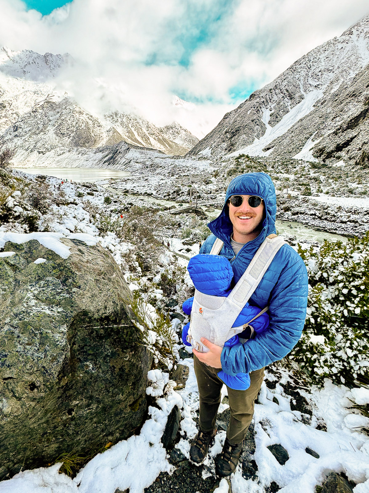 Dad holding baby in a carrier in front of a lake on the Hooker Valley Track in Mt. Cook, New Zealand