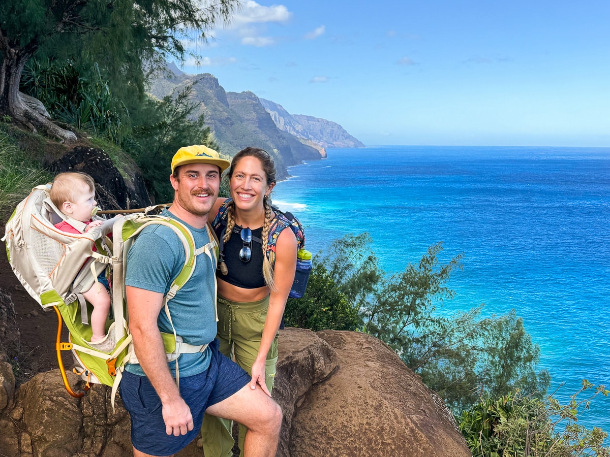 Mom, Dad, and Baby in hiking backpack stopping for a picture in front of Na Pali Coastline while hiking Hanakāpīʻai Falls Trail in Kauai, Hawaii