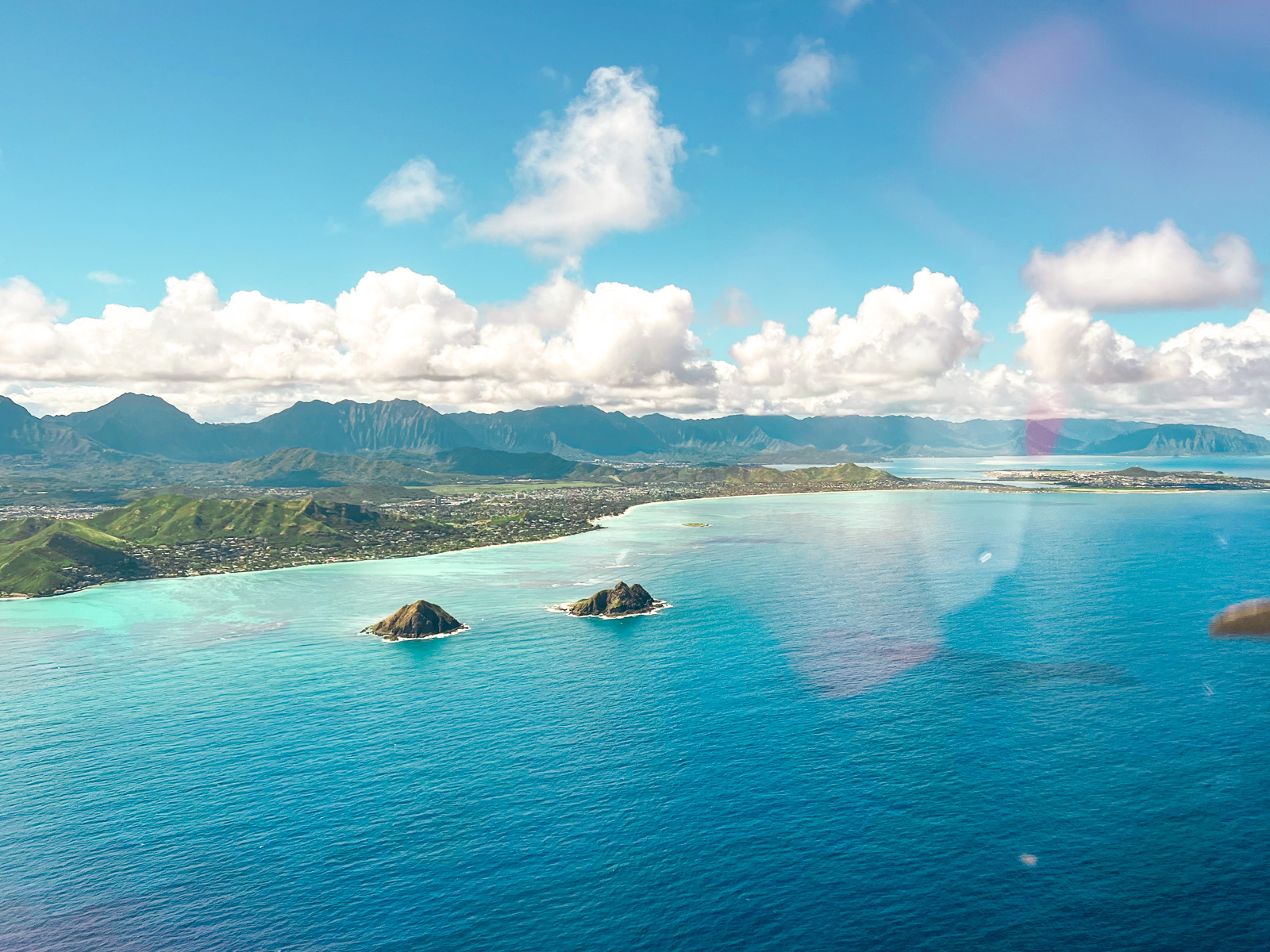 Best Spots for Whale Watching in Oahu Blog Post Picture: Aerial view of the turquoise waters and coastline on the east side of Oahu, Hawaii, with the Mokulua Islands in the foreground and lush green mountains under a partly cloudy sky