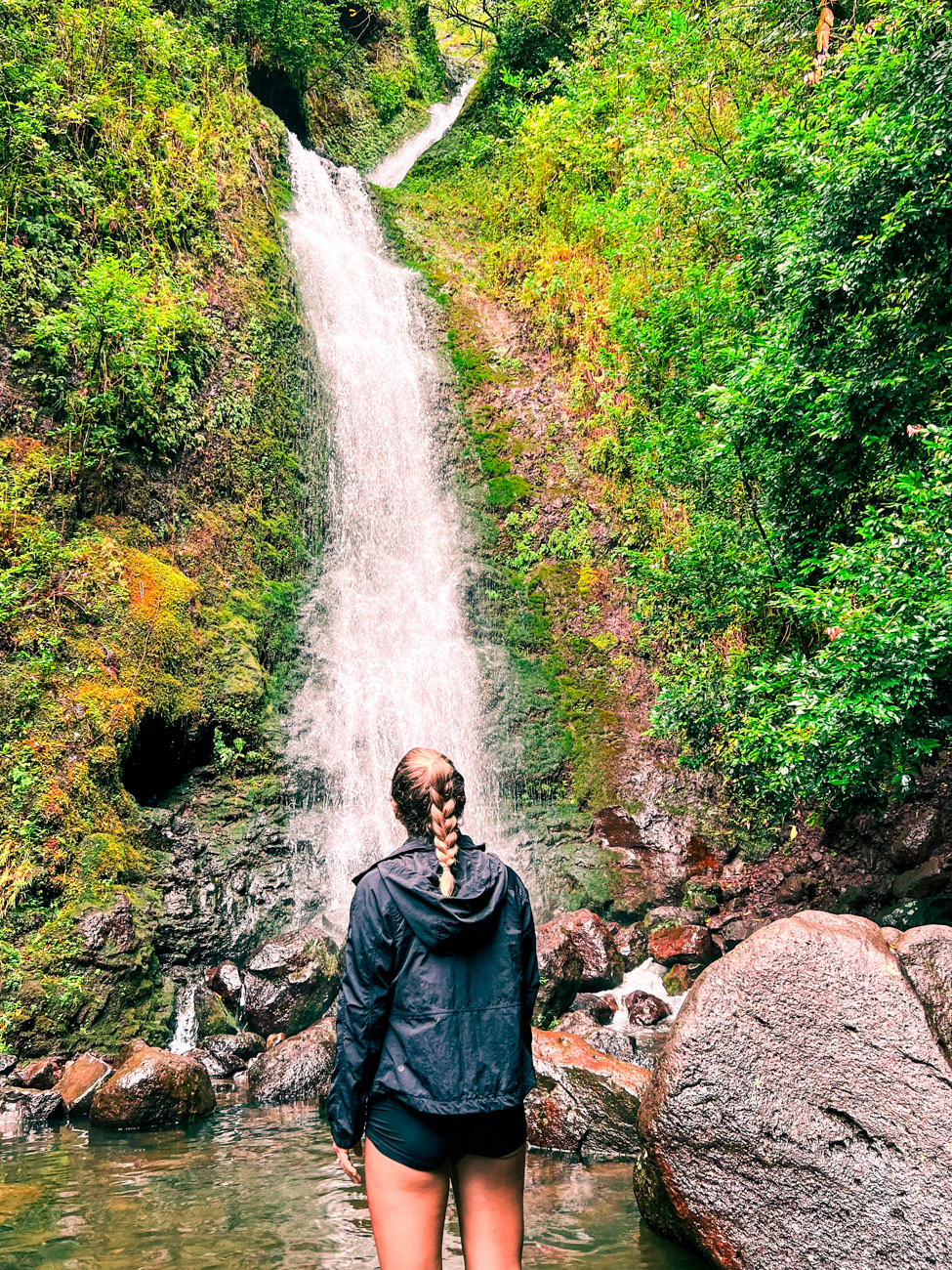 View of Lululmahu Falls in Oahu, Hawaii from below with a woman standing in the pool of the falls with rocks and boulders all around her, looking up at the waterfall in front and above her.