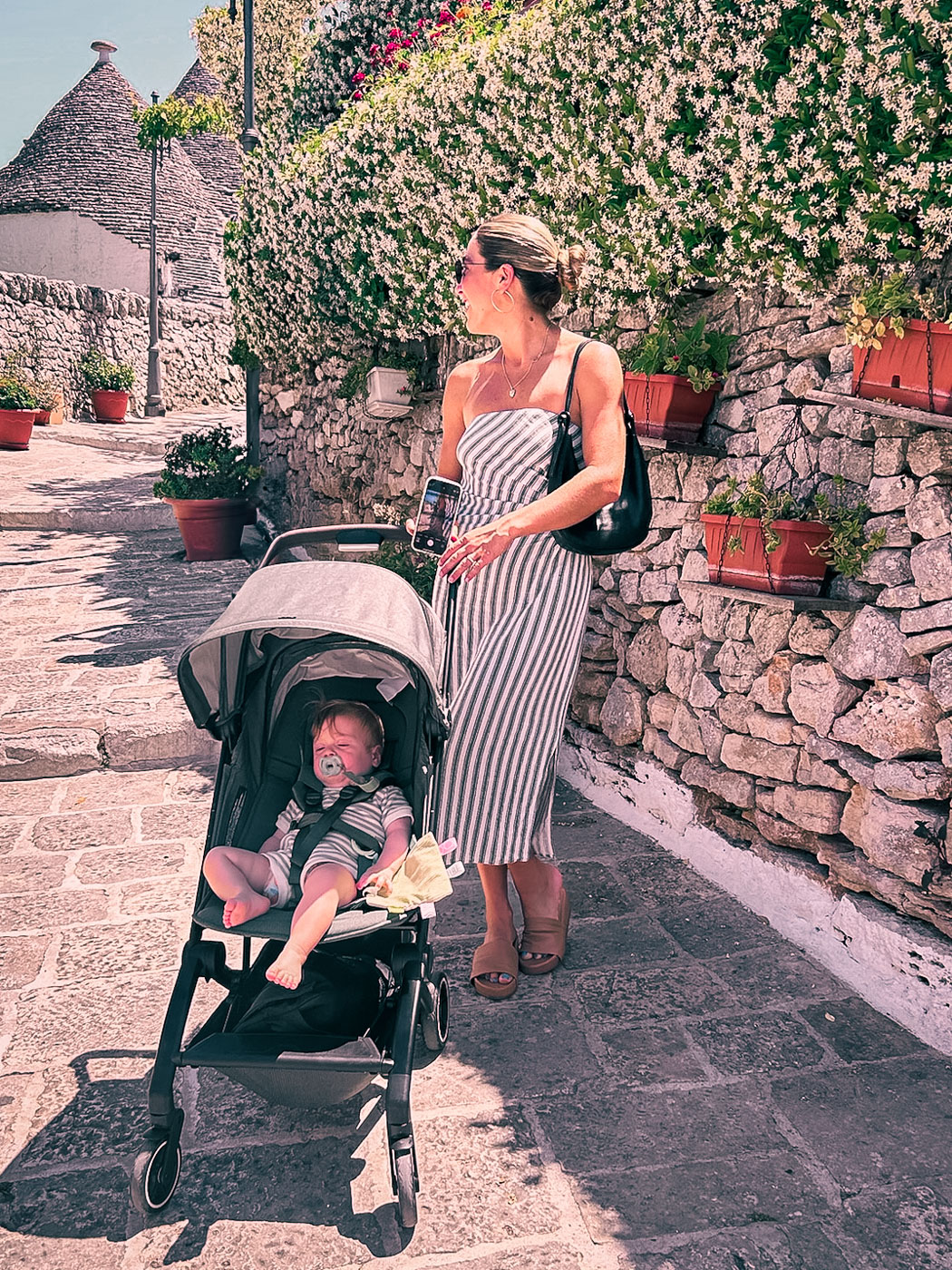Mom standing on a cobblestone street with a baby in Travel Stroller and Trulli houses in the backgroud in Alberobello, Puglia, Italy