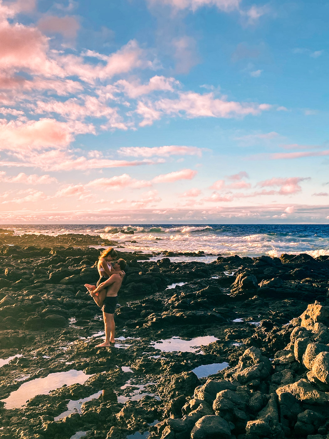Man holding woman at Ka'ena Point on the west side of Oahu, Hawaii. Rocky beach of Ka'ena Point in the foreground with the Pacific Ocean waves in the background below a mostly sunny sky