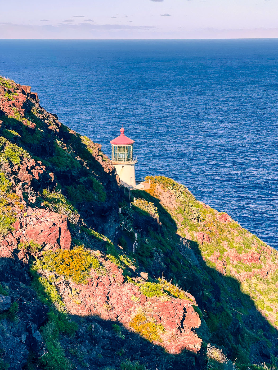 Makapu'u lighthouse as seen from Makapu'u Lighthouse Trail with the Makapu'u Point Mountainside in the foreground and the blue water of the pacific ocean in the background