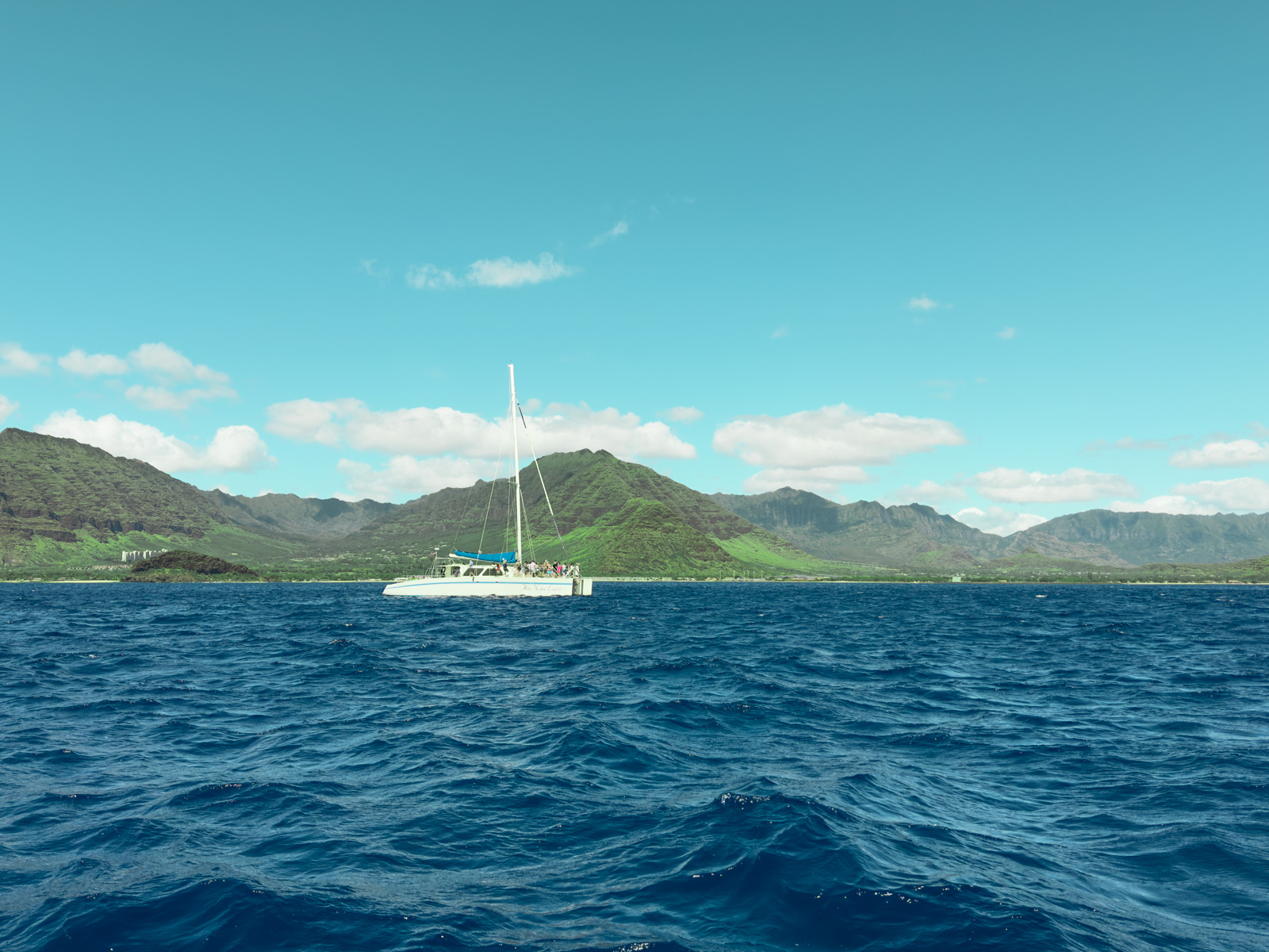 Very Best Whale Watching in Oahu Blog Post Cover Photo: A white catamaran sailboat on the ocean off the west side of Oahu, Hawaii, with a backdrop of lush green mountains under a bright blue sky with scattered clouds.