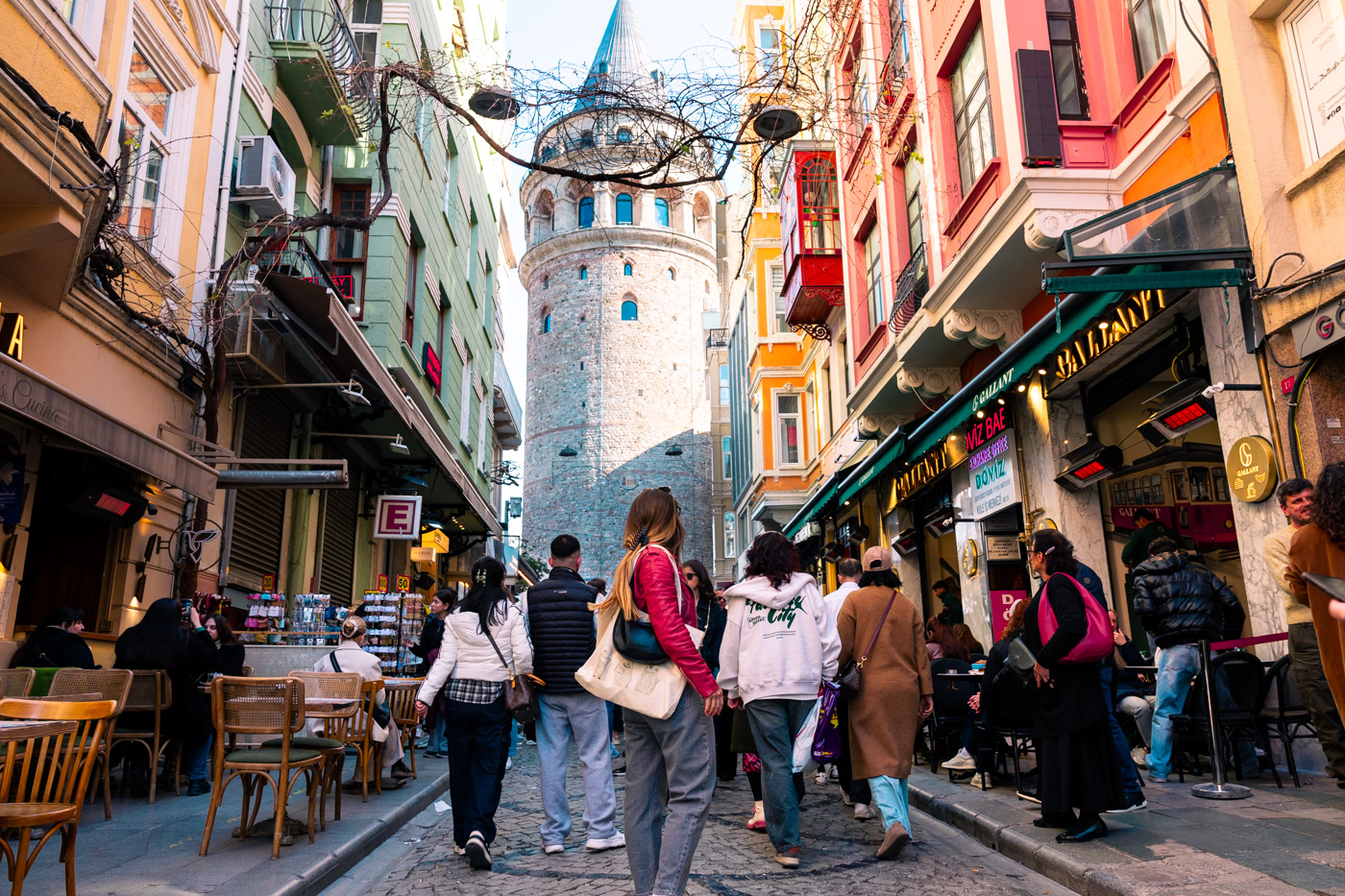 11 Best, Most Unique Things to Do in Istanbul, Turkey: A woman standing on the street in front of The Galata Tower in Istanbul Turkey with other people on the street around her and colorful shops and houses behind her.