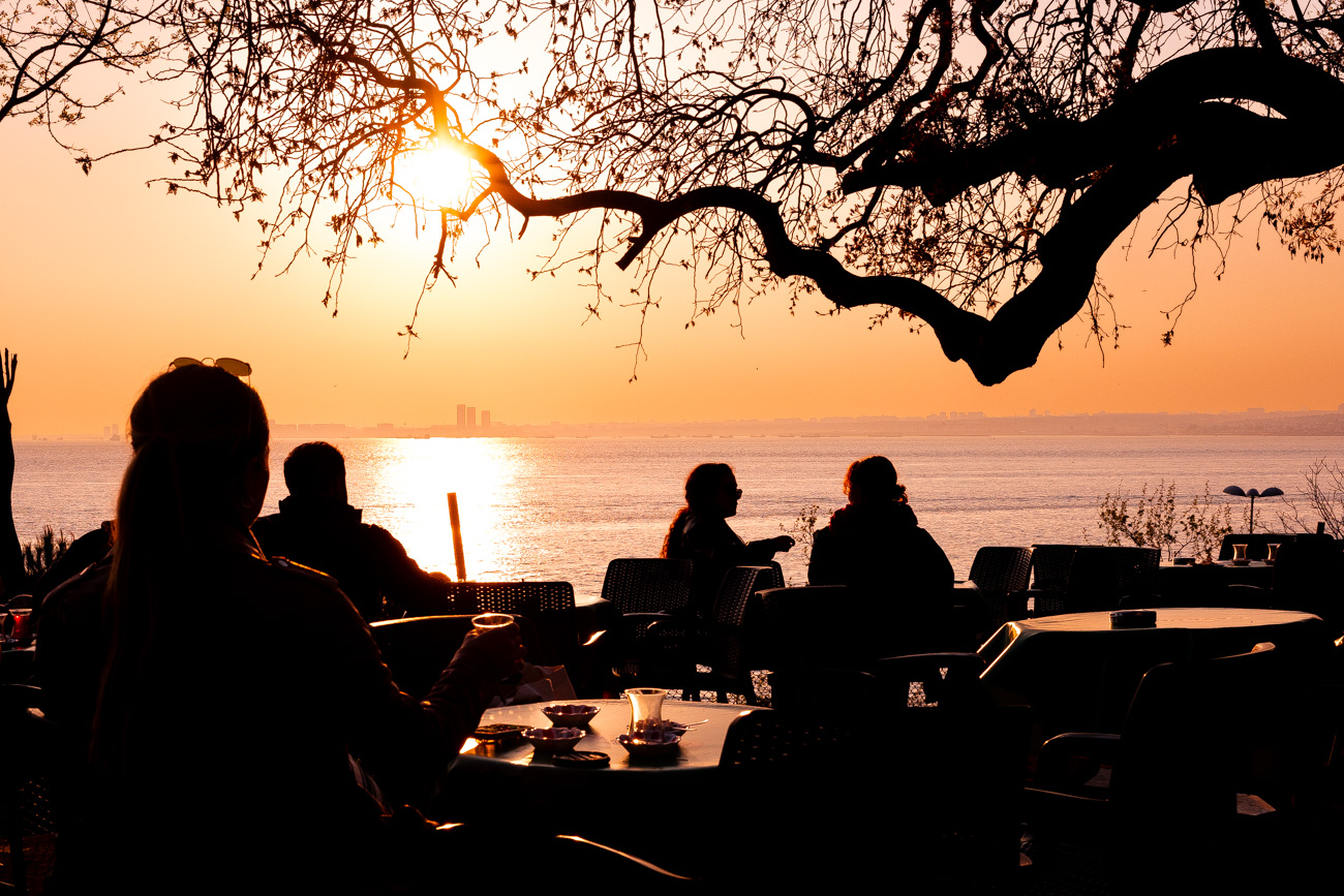 11 Best, Most Unique Things to Do in Istanbul, Turkey: A woman sitting at a table drinking tea with a tree and the sunset over the water behind her at Moda Aile Çay Bahçesi, a Family Tea Garden in Moda, Istanbul, Turkey