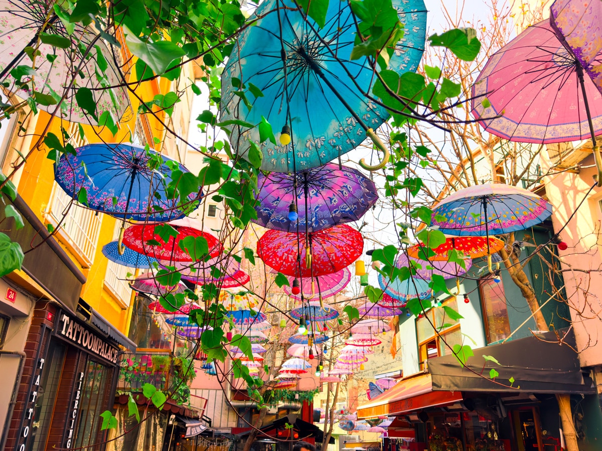 11 Best, Most Unique Things to Do in Istanbul, Turkey: Colorful umbrellas hang overhead between buildings along a narrow street in Istanbul, Turkey, creating a vibrant canopy interwoven with green vines, while shops and cafes line the lively urban scene below.